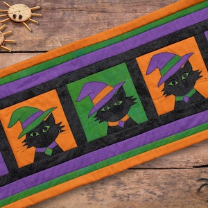May include: A Halloween-themed table runner featuring repeating panels of black cats wearing witch hats. The cats are set against orange and green backgrounds, framed by purple and orange stripes. Decorative spider-shaped snacks are on the wooden surface.
