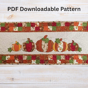 May include: A quilted table runner featuring a central panel with five appliqued pumpkins in shades of orange, red, and yellow. The pumpkins have green leaves and are set against a cream background. The border is made of patchwork squares in fall colors. The text "PDF Downloadable Pattern" is at the top.
