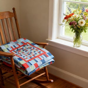 K&ouml;nnte beinhalten: Ein h&ouml;lzerner Schaukelstuhl mit einer gefalteten Steppdecke und einer Vase mit Blumen. Die Steppdecke ist blau, rot und gr&uuml;n. Die Blumen sind eine Mischung aus Farben, darunter Gelb, Orange und Lila. Der Stuhl steht in der N&auml;he eines Fensters.