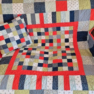 K&ouml;nnte beinhalten: Ein Patchwork-Quilt und ein passendes Kissen. Der Quilt hat einen roten Rand und ein zentrales quadratisches Design mit roten, blauen und gr&uuml;nen Quadraten. Das Kissen hat ein &auml;hnliches quadratisches Muster.