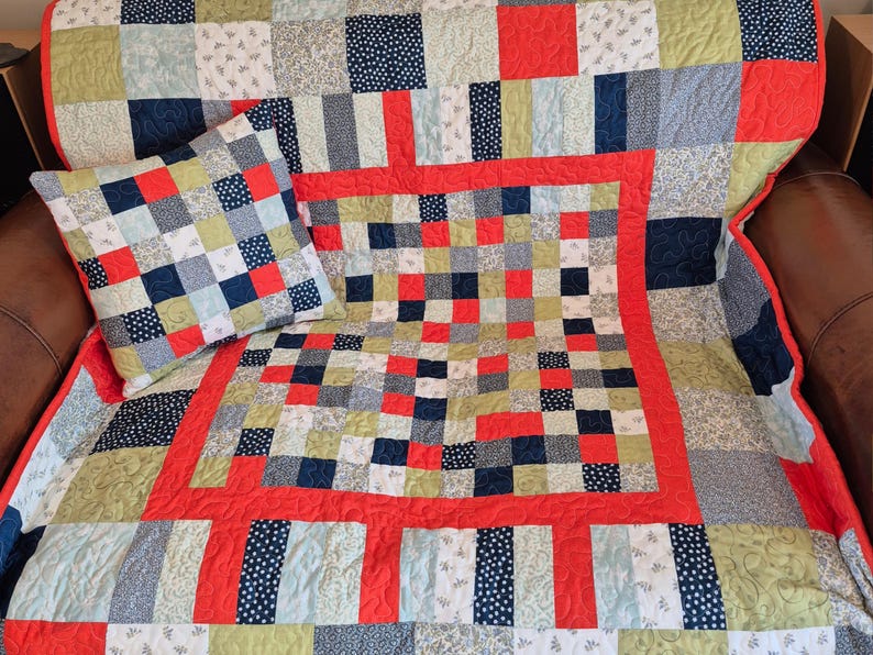 K&ouml;nnte beinhalten: Ein Patchwork-Quilt und ein passendes Kissen. Der Quilt hat einen roten Rand und ein zentrales quadratisches Design mit roten, blauen und gr&uuml;nen Quadraten. Das Kissen hat ein &auml;hnliches quadratisches Muster.