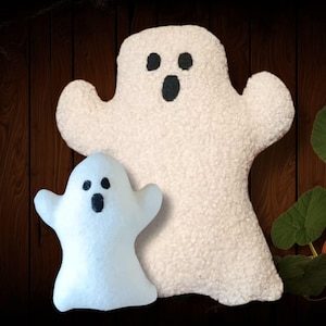 May include: Two white ghost-shaped decorations with black facial features. The larger ghost has an open mouth, and the smaller ghost has a closed mouth. The background is a wooden surface with a pumpkin and vines.