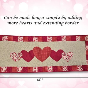 May include: A quilted table runner featuring a cream-colored center with five red hearts. The runner is 40 inches long and 14 inches wide, with a red border. The text says, "Can be made longer simply by adding more hearts and extending border."