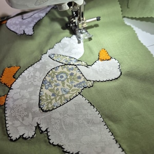 May include: A sewing machine needle is stitching a fabric applique onto a sage green background. The applique features a white floral patterned goose with an orange beak and feet, and a blue and white floral patterned wing detail.