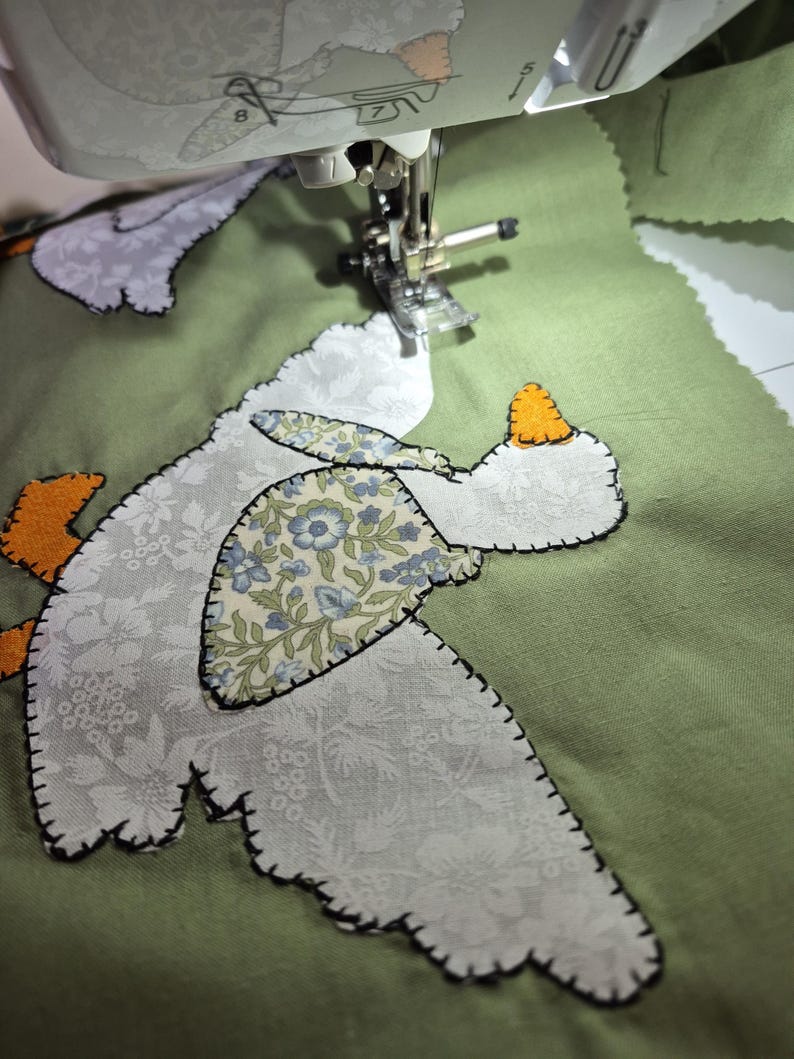 May include: A sewing machine needle is stitching a fabric applique onto a sage green background. The applique features a white floral patterned goose with an orange beak and feet, and a blue and white floral patterned wing detail.