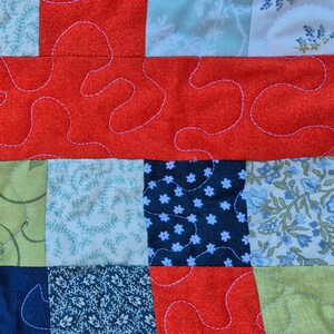 K&ouml;nnte beinhalten: Nahaufnahme eines Patchwork-Quilts mit einer Vielzahl von gemusterten Stoffen. Der Quilt zeigt Quadrate aus floralen, Blatt- und unifarbenen Stoffen in Blau-, Gr&uuml;n- und Rott&ouml;nen. Wei&szlig;es Garn erzeugt ein dekoratives, wellenf&ouml;rmiges Muster.
