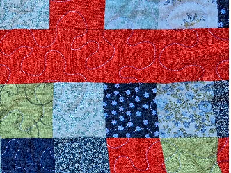 K&ouml;nnte beinhalten: Nahaufnahme eines Patchwork-Quilts mit einer Vielzahl von gemusterten Stoffen. Der Quilt zeigt Quadrate aus floralen, Blatt- und unifarbenen Stoffen in Blau-, Gr&uuml;n- und Rott&ouml;nen. Wei&szlig;es Garn erzeugt ein dekoratives, wellenf&ouml;rmiges Muster.