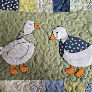 May include: A quilted fabric panel featuring two white geese appliqu&eacute;s on a sage green background. The left goose wears a patterned neck scarf, while the right goose wears a dark blue floral neck scarf. Both geese have orange feet and beaks.