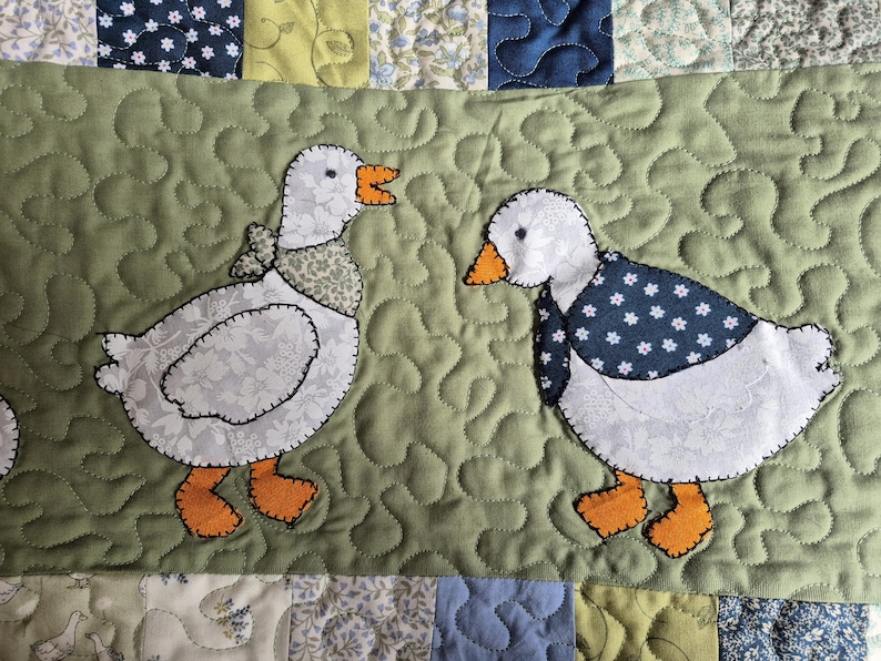 May include: A quilted fabric panel featuring two white geese appliqu&eacute;s on a sage green background. The left goose wears a patterned neck scarf, while the right goose wears a dark blue floral neck scarf. Both geese have orange feet and beaks.