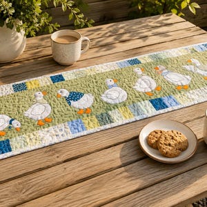 May include: A rectangular quilted table runner featuring a light green center panel with an appliqued row of six white geese wearing blue and white patterned accessories. The border consists of a patchwork of blue, yellow, and white squares.