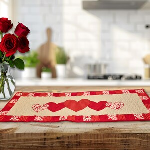 May include: A rectangular table runner with a cream-colored center and a red border. The runner features three red hearts and two hearts with a white and red pattern. A vase of red roses sits to the left.