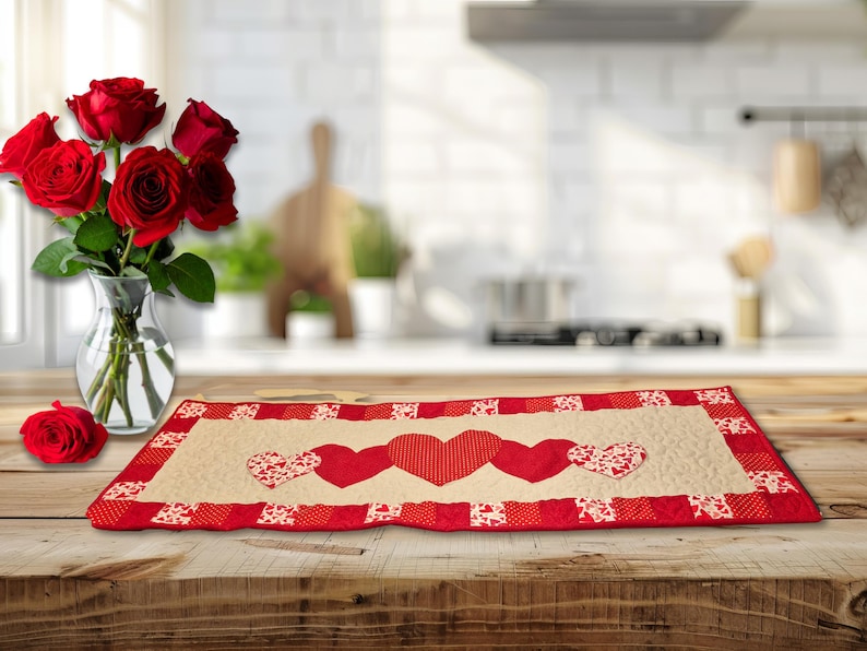 May include: A rectangular table runner with a cream-colored center and a red border. The runner features three red hearts and two hearts with a white and red pattern. A vase of red roses sits to the left.