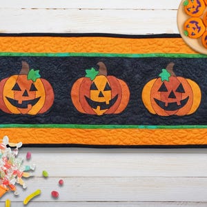 May include: A Halloween table runner with three smiling pumpkin faces on a black background, framed by orange and green borders. The image also shows orange cookies and a pumpkin-shaped candy bucket.