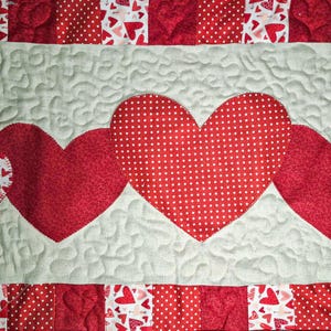 May include: A quilted textile featuring a design of red hearts on a light gray background. The hearts are of varying sizes and patterns, including polka dots and solid red. The quilt has a patchwork border with heart and polka dot patterns.