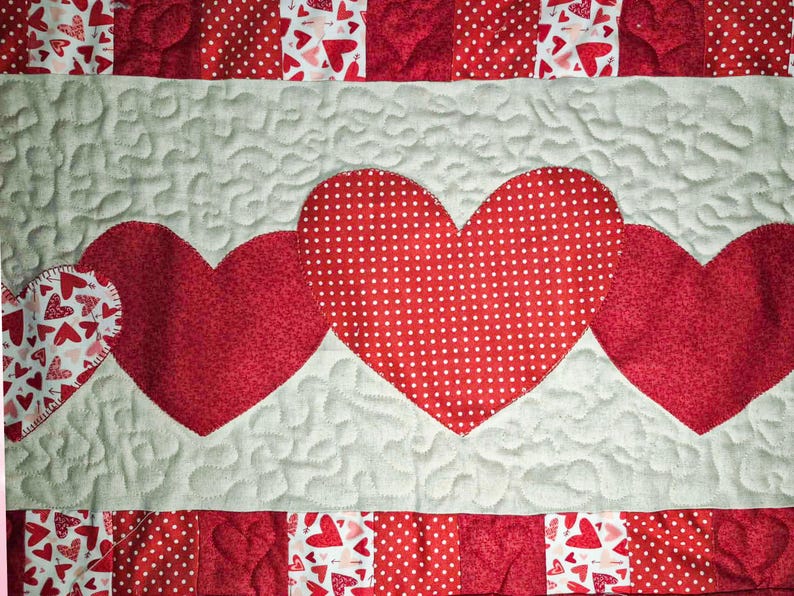 May include: A quilted textile featuring a design of red hearts on a light gray background. The hearts are of varying sizes and patterns, including polka dots and solid red. The quilt has a patchwork border with heart and polka dot patterns.