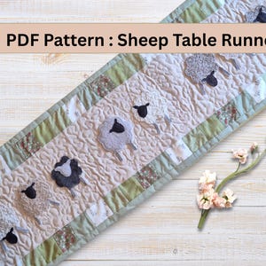 Könnte beinhalten: Gesteppter Tischläufer mit Schafmuster. Der Läufer hat einen hellbeigen Hintergrund mit welliger Textur, umrandet von grünem und floralem Stoff. Der Text "PDF Pattern : Sheep Table Runner" befindet sich oben. Er ist ca. 150 cm lang.