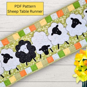 Peut inclure: Chemin de table matelassé avec des appliqués de moutons. Le fond est vert avec des moutons blancs et des visages noirs. La bordure est composée de carrés orange, verts et blancs. Le texte de l'image indique "PDF Pattern Sheep Table Runner."