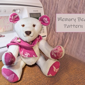 Könnte beinhalten: Ein handgefertigter weißer Teddybär mit rosa floralen Akzenten an Ohren, Schal und Pfoten. Der Bär hat braune Augen und eine genähte Nase. Der Text "Memory Bear Pattern" wird auf einem rosa Rechteck angezeigt. Im Hintergrund ist eine Nähmaschine zu sehen.