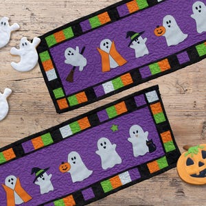以下が含まれることがあります： 紫色の中心部と幽霊とカボチャのデザインが施された、ハロウィーンをテーマにした2つのテーブルランナー。 ランナーは黒、緑、オレンジの正方形で縁取られています。 3つの幽霊の形をしたクッキーとカボチャのクッキーも存在します。