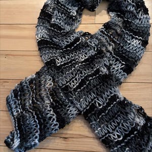 Pode incluir: Um cachecol de malha à mão em tons de cinza, preto e branco. O cachecol é feito com um ponto texturizado, criando um padrão visualmente interessante. O cachecol está sobre uma superfície de madeira.