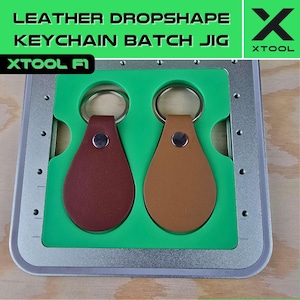Puede incluir: Un jig de metal plateado con dos llaveros de cuero verde. Los llaveros tienen forma de lágrima y un anillo de metal plateado. El jig está etiquetado como "Leather Dropshape Keychain Batch Jig" y "XTOOL F1".