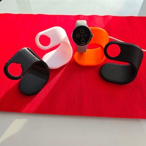 Può includere: Tre supporti per orologi stampati in 3D in nero, bianco e arancione. I supporti sono progettati per contenere uno smartwatch con quadrante rotondo.