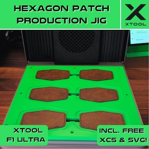 May include: A green production jig with six hexagon-shaped cutouts for leather patches. The jig is labeled "HEXAGON PATCH PRODUCTION JIG" and "XTOOL F1 ULTRA". The jig is sitting on a wooden surface. The image also includes the text "INCL. FREE XCS & SVG!"