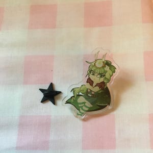 May include: A clear acrylic charm featuring a green and brown cartoon character with a star-shaped pin. The character has green skin, a crown, and is sitting down. The star pin is black.