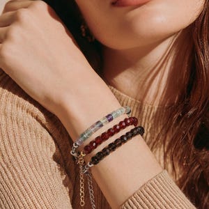 May include: Three beaded bracelets in shades of green, red, and brown adorn a wrist. The bracelets feature small, faceted beads strung on a chain. The wrist is against a neutral background, with the brand name "arvin" visible in the bottom right corner.