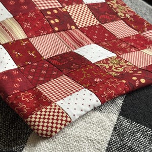 Pode incluir: Uma bolsa de tecido retangular com um design de patchwork composto por vários quadrados vermelhos, brancos e creme. Os quadrados de tecido exibem diversos padrões, incluindo estampas florais, xadrez e geométricas. Um zíper bege é visível no topo.