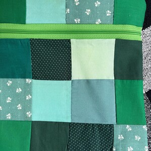 Puede incluir: Una bolsa de tela rectangular con un diseño de patchwork en varios tonos de verde. Los cuadrados incluyen verde sólido, lunares blancos sobre verde oscuro y patrones de mariposas blancas sobre verde claro. Una cremallera verde brillante en la parte superior.