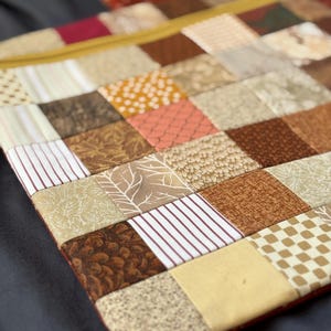 May include: A square fabric pouch featuring a patchwork design of various brown, tan, and cream-coloured squares. The patterns include stripes, dots, checks, and botanical prints. A mustard yellow zip runs along the top edge of the pouch.