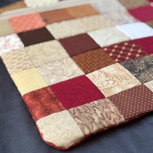 May include: A rectangular zipped pouch constructed from a patchwork of fabric squares in various shades of brown, cream, burgundy, and tan. The textile features a mix of solid colours and diverse patterns, including dots, swirls, and geometric prints.