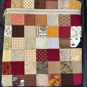 May include: A rectangular fabric pouch featuring a patchwork design of various square fabric swatches in shades of red, brown, tan, and cream. The pouch includes a beige zip closure along the top edge and displays diverse patterns including skulls, leaves, checks, and a face with red lips.