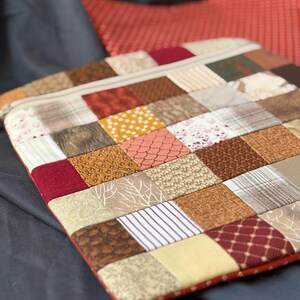 Könnte beinhalten: Eine quadratische Stofftasche mit einem Patchwork-Design aus verschiedenen kleinen Quadraten in Braun-, Beige-, Creme- und Dunkelrottönen. Die Muster umfassen Streifen, Punkte, florale Motive und geometrische Formen. Ein Reißverschluss ist oben sichtbar.