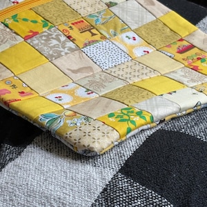 Könnte beinhalten: Eine quadratische Reißverschlusstasche aus einem Patchwork aus Stoffquadraten in verschiedenen Gelb-, Creme- und Weißtönen. Der Stoff zeigt diverse Muster wie Kirschen, florale Motive und kleine Hasen, abgerundet durch einen gelben Reißverschluss.