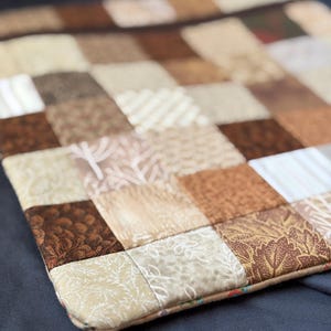Puede incluir: Una alfombrilla de tela acolchada cuadrada con un diseño de patchwork de varios cuadrados en tonos marrón, beige y crema. Los cuadrados individuales muestran diversos patrones, incluyendo motivos de hojas, rayas finas y texturas lisas.