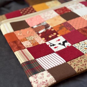 May include: A rectangular zipped pouch featuring a patchwork design composed of various fabric squares in shades of red, brown, orange, and cream. The patterns include stripes, plaids, floral prints, and a small section with skull motifs.