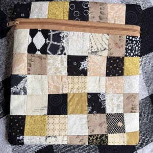 May include: A square zippered pouch constructed from a patchwork of fabric squares in black, white, cream, and mustard yellow. The fabric features various patterns including floral, geometric, and polka dot prints. A tan zip runs across the top.