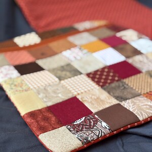 Puede incluir: Una funda de cojín cuadrada de patchwork que presenta una cuadrícula de varios cuadrados de tela en tonos rojo, marrón, beige y crema. Los cuadrados textiles muestran diversos patrones, incluyendo texturas geométricas, florales y lisas, con borde rojo.