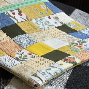 Pode incluir: Uma bolsa de tecido retangular com um design de patchwork de vários quadrados de algodão em tons de amarelo, verde-oliva, azul e branco. A superfície exibe padrões florais, de morangos e geométricos. Um zíper verde-claro percorre a borda superior.