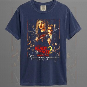 May include: A navy blue short-sleeve t-shirt featuring a graphic print of the movie poster for Ready or Not 2. The design includes multiple characters, the title text Ready or Not 2, and the phrase Here I Come in bold red and yellow lettering.