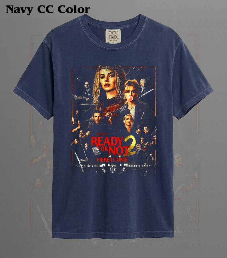 May include: A navy blue short-sleeve t-shirt featuring a graphic print of the movie poster for Ready or Not 2. The design includes multiple characters, the title text Ready or Not 2, and the phrase Here I Come in bold red and yellow lettering.