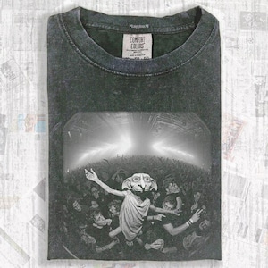 Può includere: T-shirt Comfort Colors grigio scuro con una grafica in bianco e nero di una folla e un personaggio. La maglietta ha girocollo e maniche corte. L'etichetta recita "Comfort Colors".