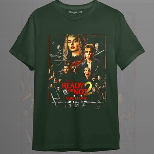 May include: A forest green short-sleeve t-shirt featuring a graphic print of the movie poster Ready or Not 2 Here I Come. The design includes multiple character portraits in a dark, cinematic style with red and yellow text accents on the front.