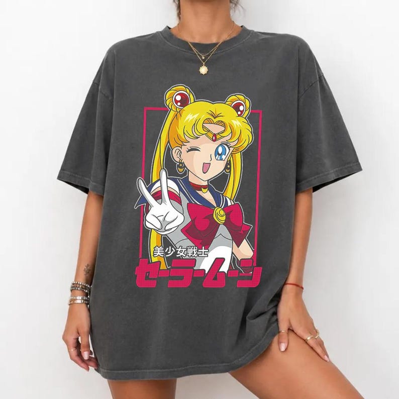 May include: A charcoal gray short-sleeve t-shirt featuring a graphic print of the anime character Sailor Moon with blonde hair, a blue and white sailor collar, and a red bow. The design includes a pink rectangular border and Japanese text.