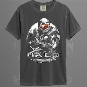 Könnte beinhalten: Ein graphitfarbenes Kurzarm-T-Shirt mit einem Grafikdruck eines futuristischen gepanzerten Soldaten in einem Helm mit orangefarbenem Visier. Das Design enthält den Text Halo Combat Evolved in weißer Schrift unterhalb der Figur.