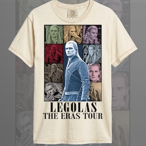May include: Ivory-colored t-shirt featuring a graphic of Legolas from The Lord of the Rings. The design includes multiple images of the character and the text "LEGOLAS THE ERAS TOUR" in a bold font. The shirt is made of a soft material.