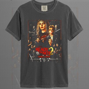 May include: A graphite-colored short-sleeve t-shirt featuring a graphic print of the movie poster for Ready or Not 2. The design includes multiple character portraits in a dark, cinematic style with the red and yellow text Ready or Not 2 Here I Come.