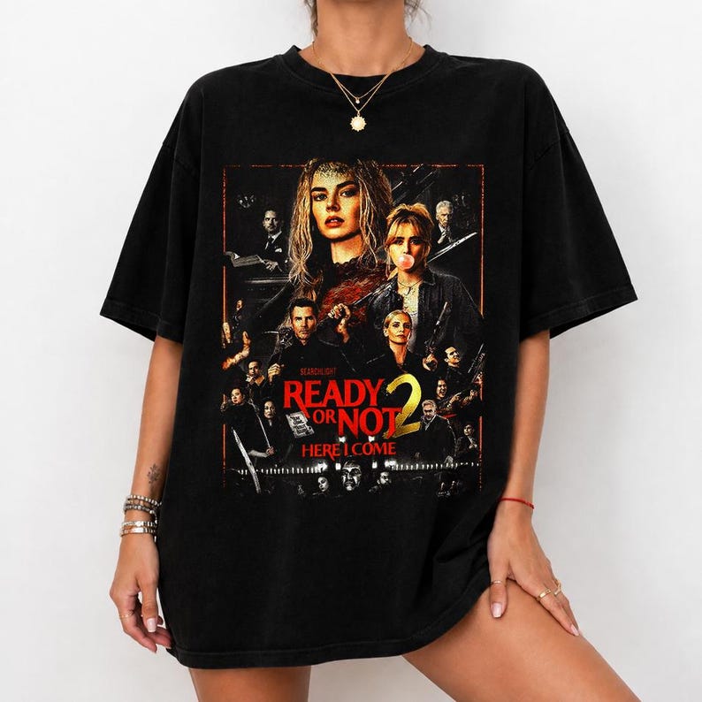 May include: A black short-sleeve t-shirt featuring a large graphic print on the front. The design displays a movie poster for Ready or Not 2, Here I Come, with multiple character portraits in dark tones and bold red text against a black background.
